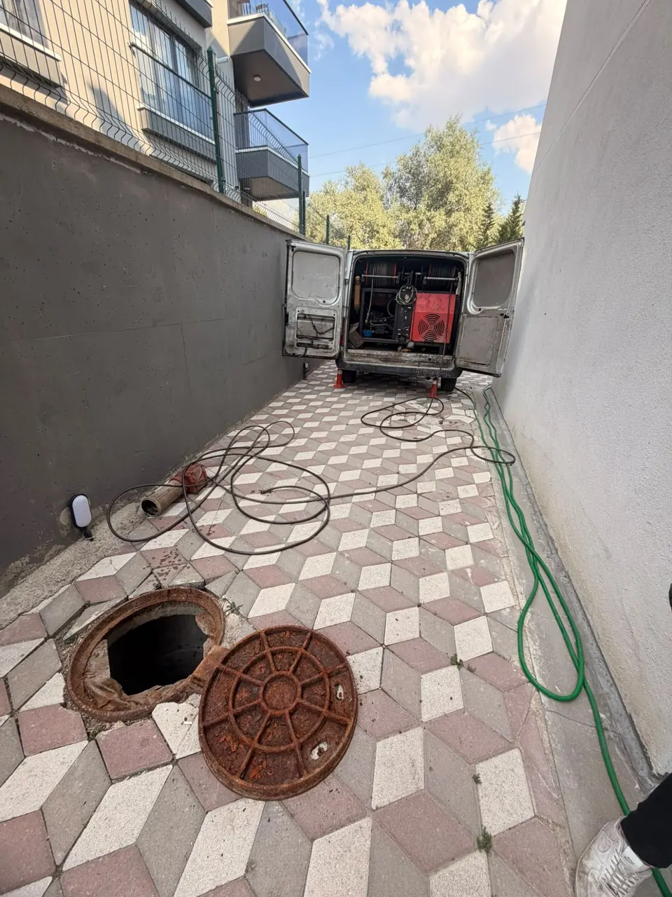 Dar bir site geçidinde açılmış rögar kapağı, araçta hortum makaraları ve pompa